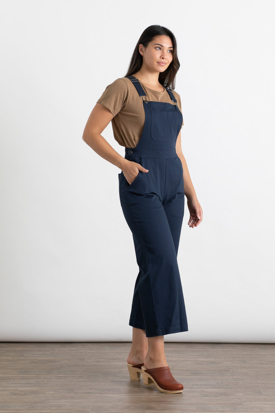 A woman stands on a wood floor against a white background, wearing Bridge & Burn's Roscoe Overall in navy canvas with a brown T-shirt and clogs, one hand in her pocket and a slight smile on her face.