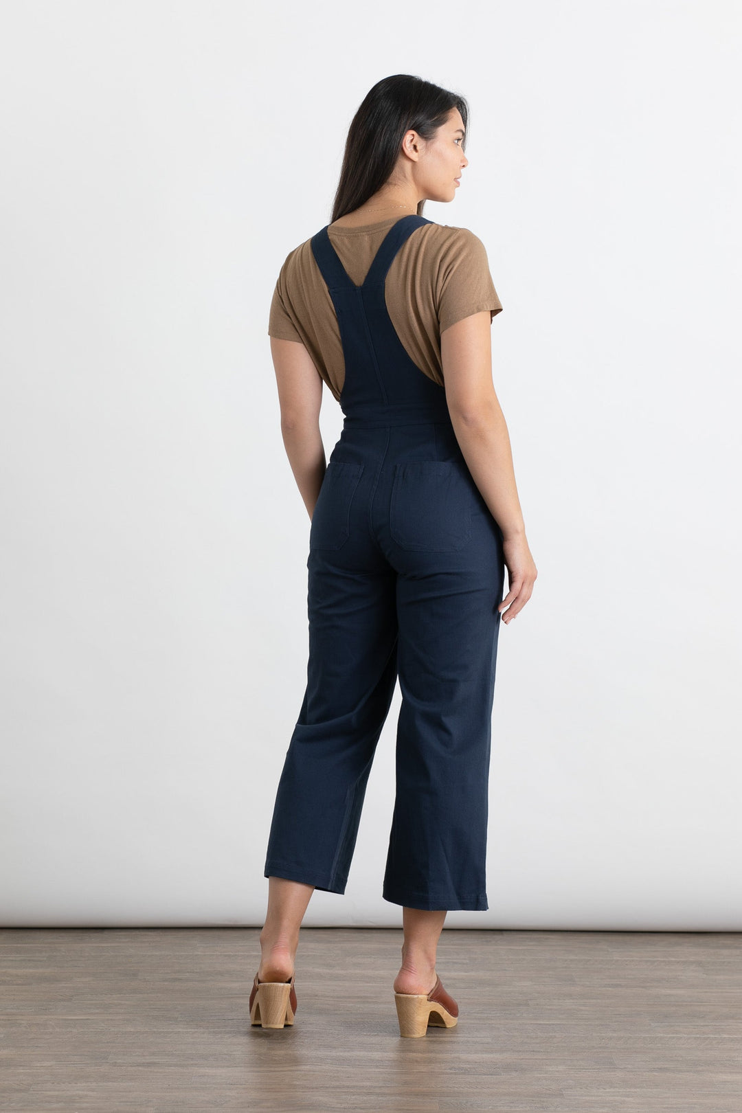 A woman faces away in Bridge & Burn’s Roscoe Overall—navy, high-waisted, wide-leg, cotton canvas—over a brown short-sleeve shirt. She wears tan heeled sandals on a plain white background with a wooden floor.