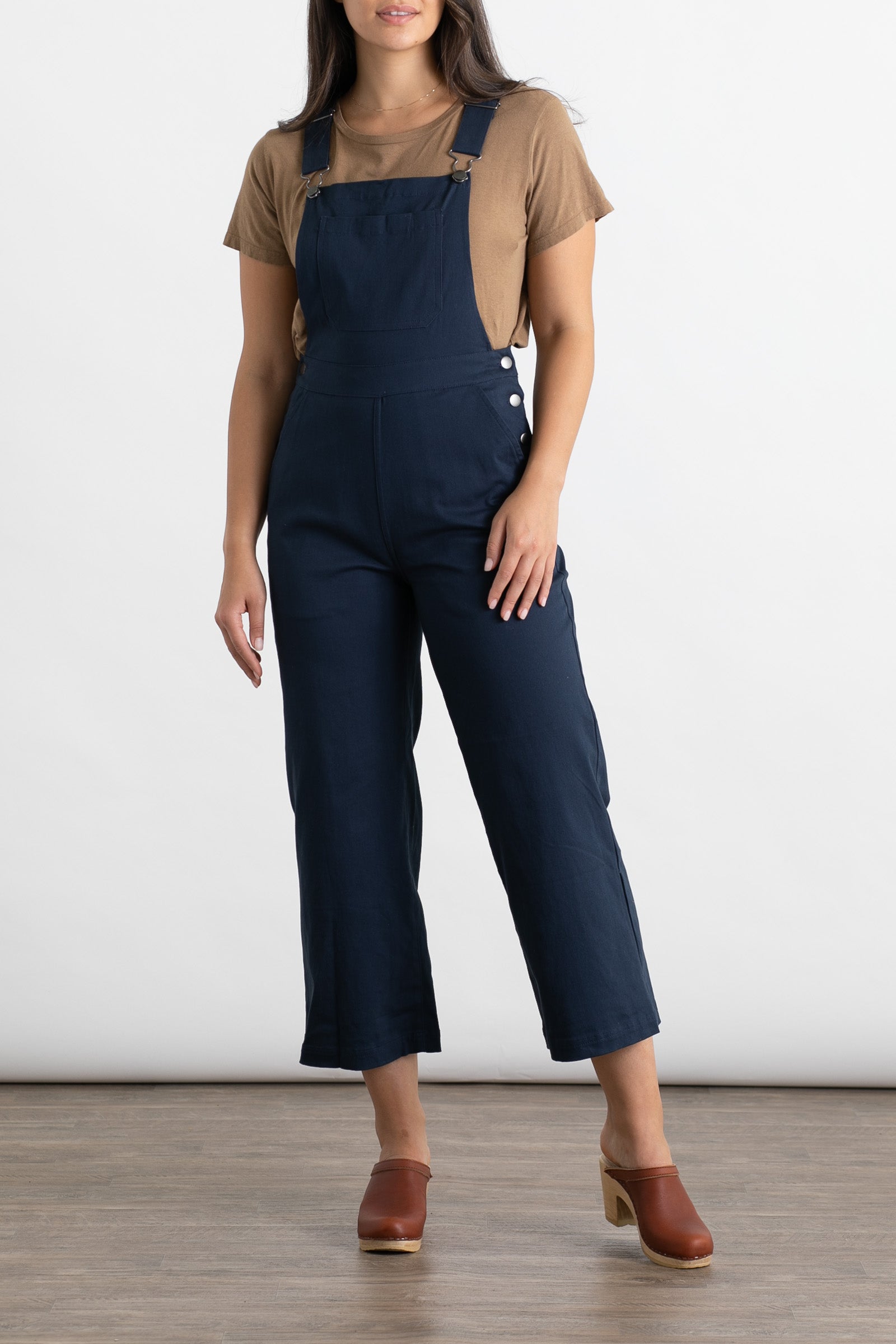 A person models Bridge & Burn’s Roscoe Overall in navy canvas, paired with a tan short-sleeve shirt and brown heeled clogs, standing on a wood floor against a plain white background.