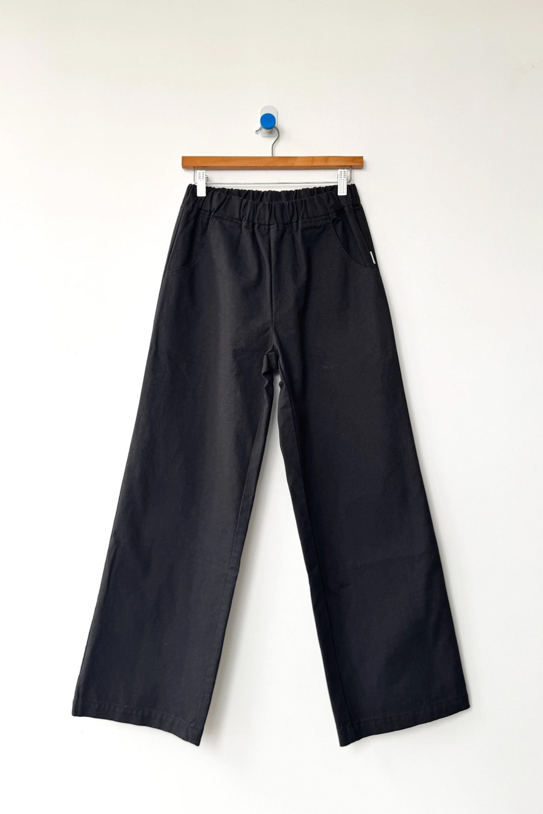A pair of Le Bon Shoppe - Work Pant / Black, crafted from heavyweight canvas with an elastic waistband, hangs on a wooden hanger against a plain white wall.