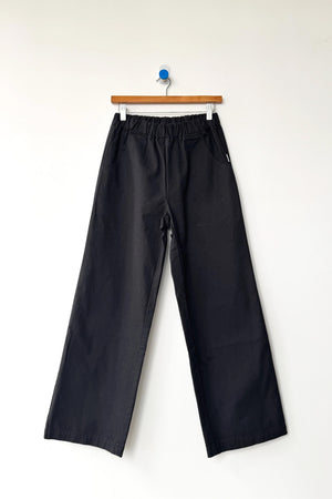 A pair of Le Bon Shoppe - Work Pant / Black, crafted from heavyweight canvas with an elastic waistband, hangs on a wooden hanger against a plain white wall.