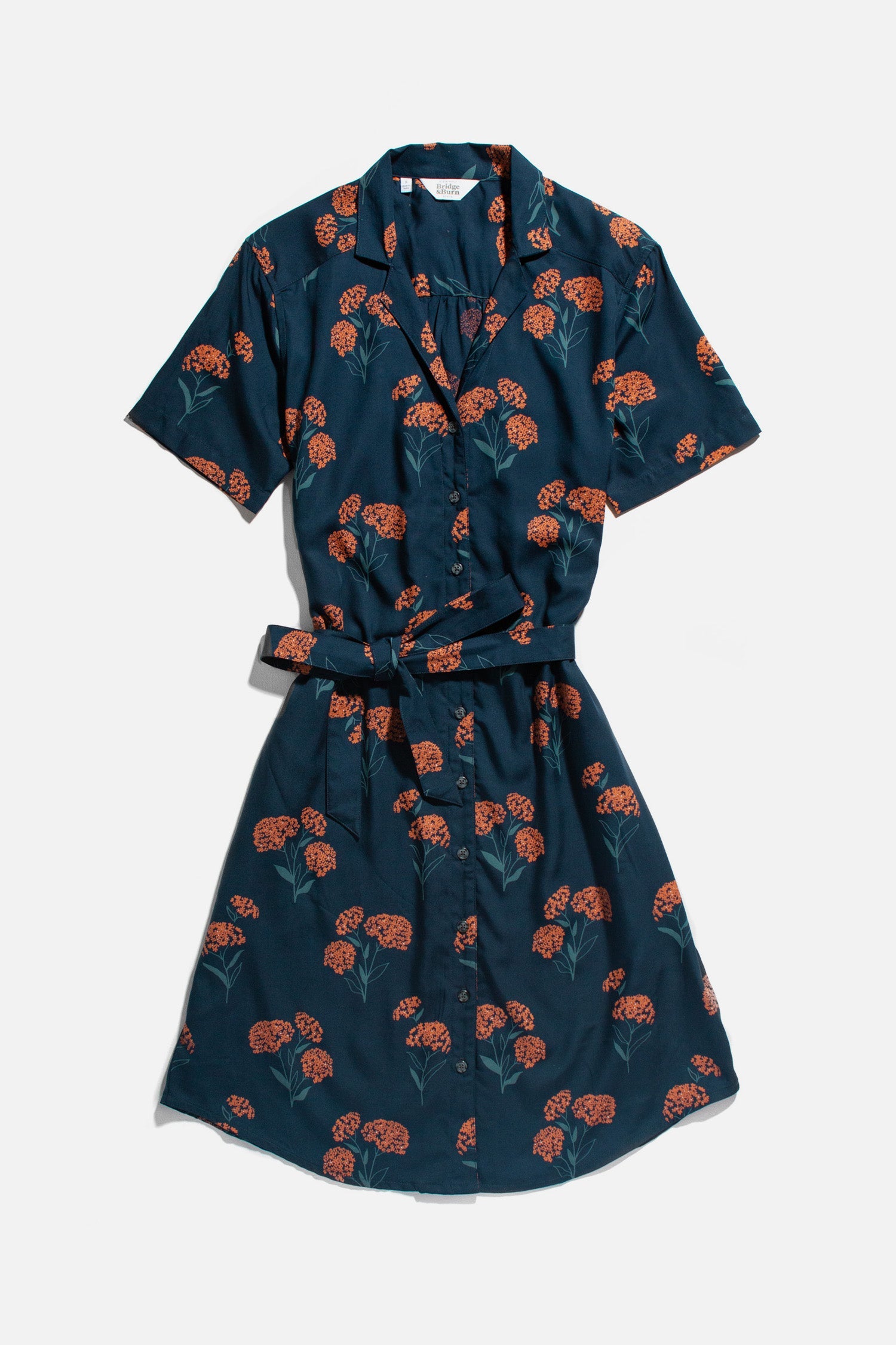 The Sofia Shirt Dress / Wildflower by Bridge & Burn is a navy blue, short-sleeved, knee-length modal dress with a collar, button front, self-belt, and orange floral pattern—ideal for sustainable summer style.