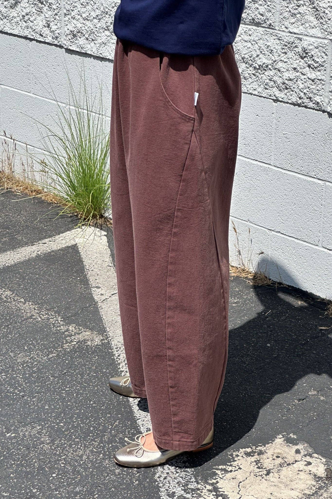 A person stands outdoors on pavement near a white brick wall, wearing the Le Bon Shoppe - Arc Pant in Chocolate with a dark blue top and silver ballet flats. Grass grows through cracks near the wall.
