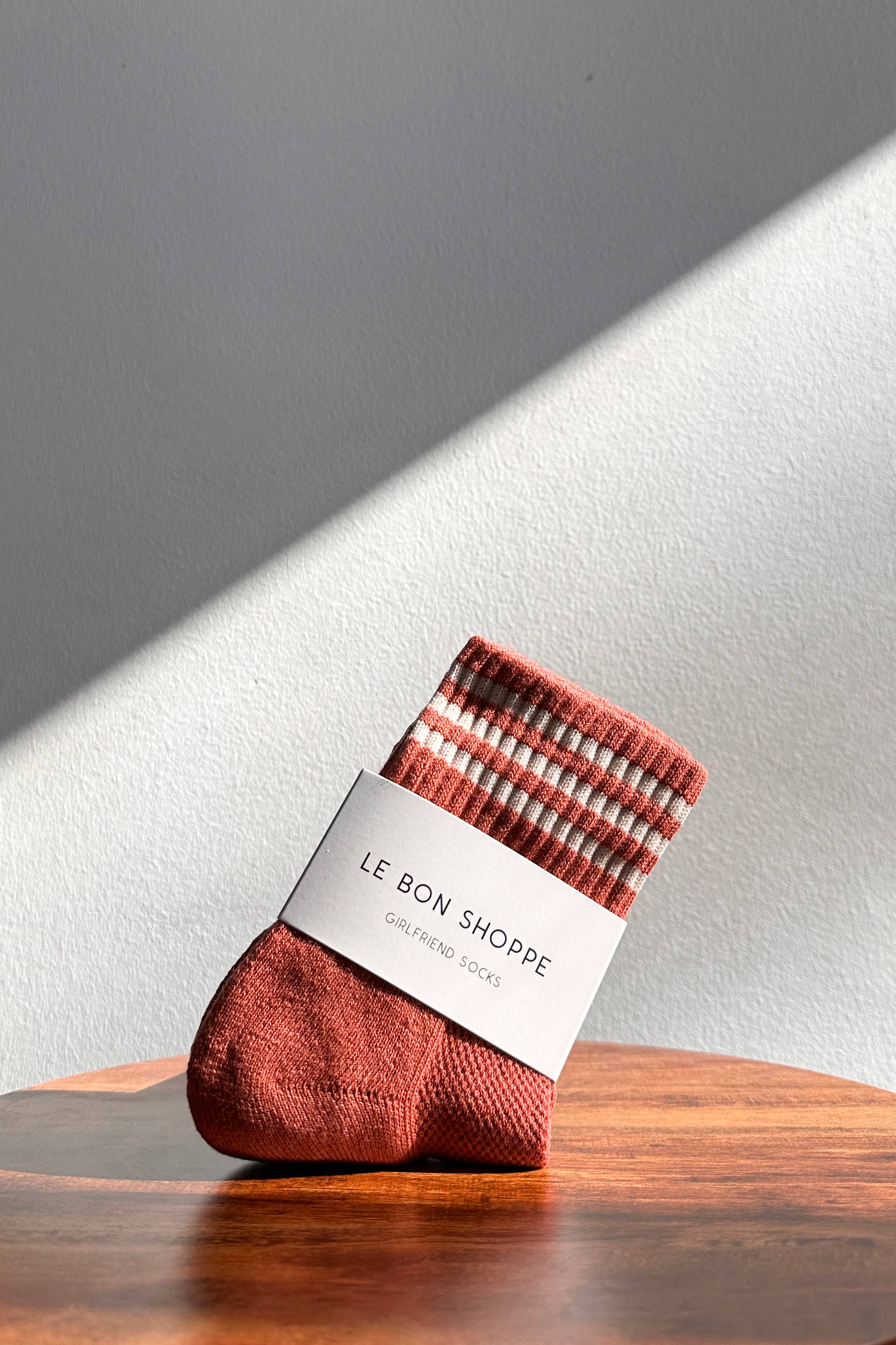 A pair of Le Bon Shoppe’s Girlfriend Socks in Terracotta, featuring white stripes, rests on a wooden surface with sunlight casting a diagonal shadow across a plain wall.