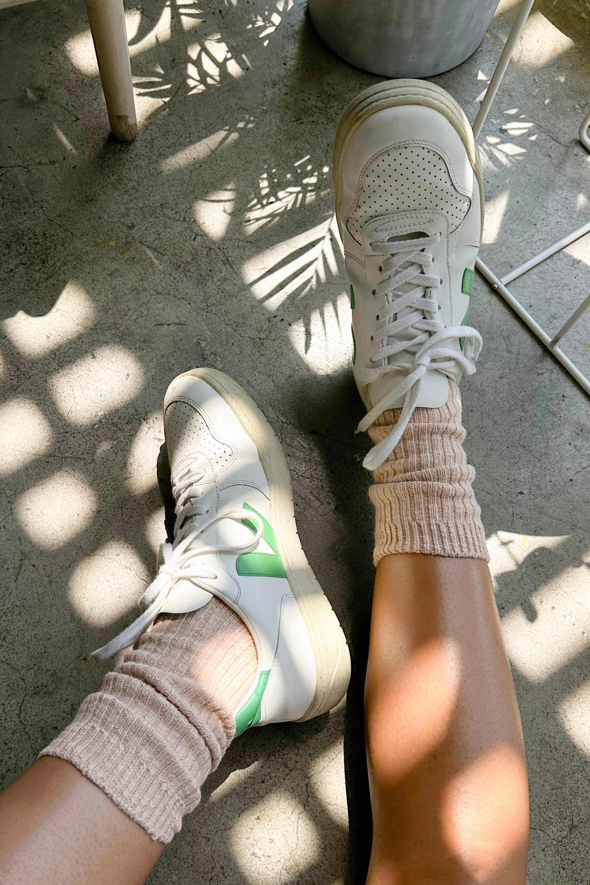 Close-up of a person wearing white sneakers and Le Bon Shoppe - Cottage Socks / Peachy Keen, sitting on a sunlit concrete floor with plant shadows and part of a white chair visible.