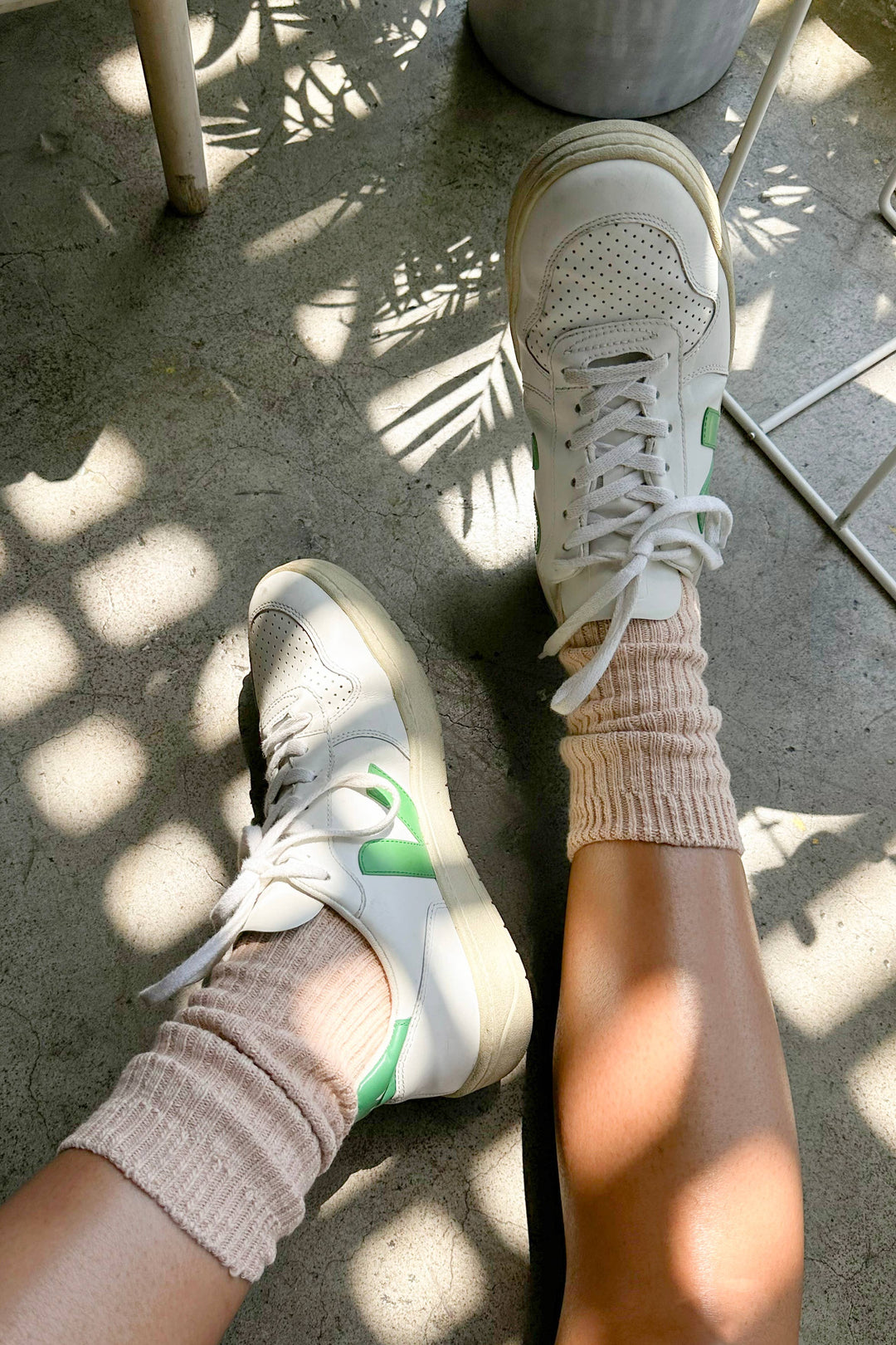 Close-up of a person wearing white sneakers and Le Bon Shoppe - Cottage Socks / Peachy Keen, sitting on a sunlit concrete floor with plant shadows and part of a white chair visible.