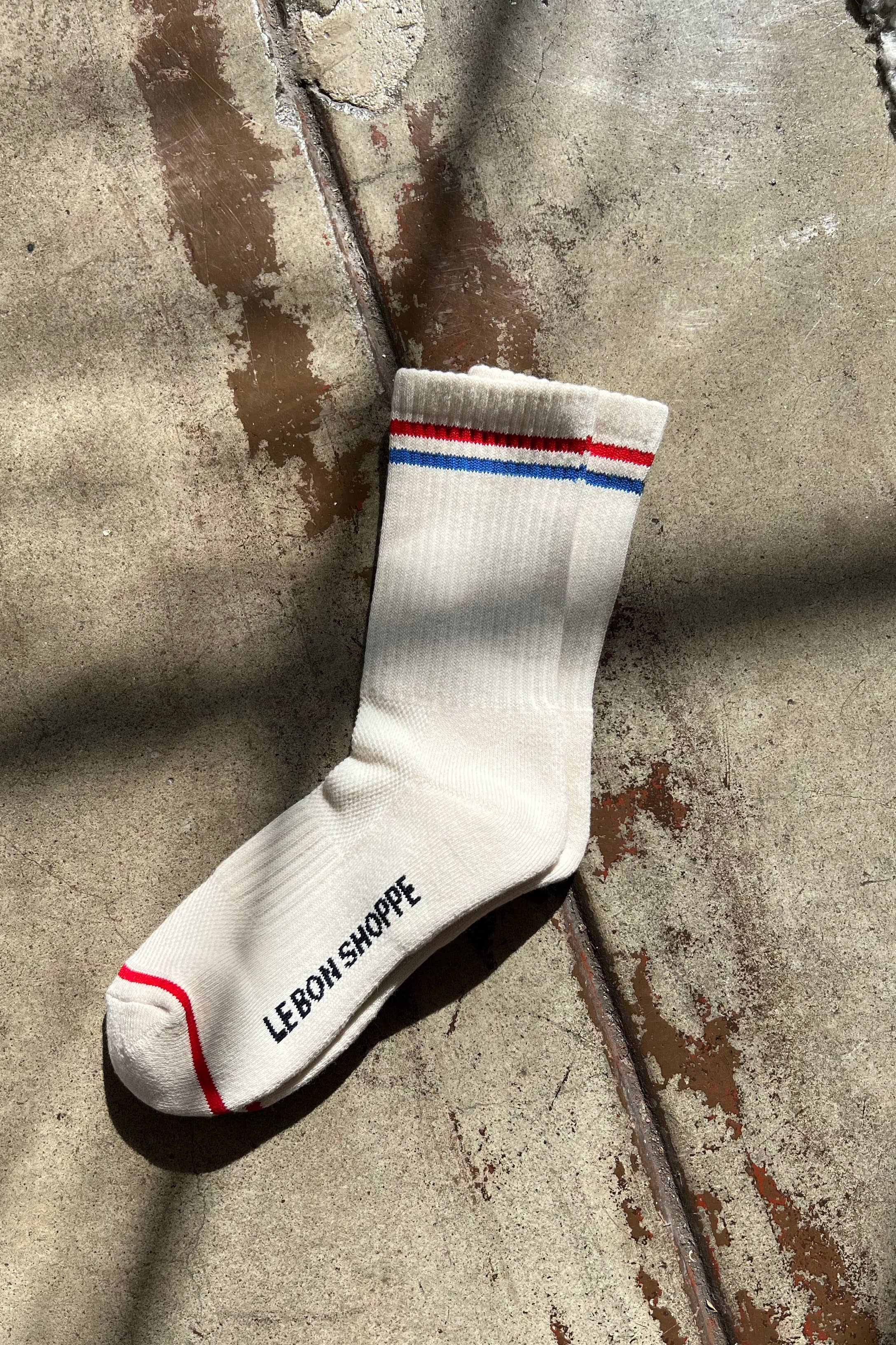The Le Bon Shoppe - Boyfriend Socks in Milk features red and blue stripes at the top, a red toe stripe, and "LE BON SHOPPE" printed in black on the sole. The cream-colored sock is shown on a sunlit, textured concrete floor.