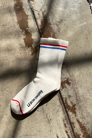 The Le Bon Shoppe - Boyfriend Socks in Milk features red and blue stripes at the top, a red toe stripe, and "LE BON SHOPPE" printed in black on the sole. The cream-colored sock is shown on a sunlit, textured concrete floor.