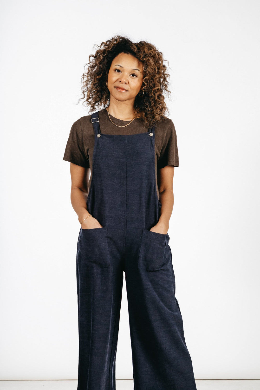 A person with curly hair wearing a brown t-shirt and the Bridge & Burn Edie Overall in Navy, featuring adjustable straps and wide legs, stands with hands in pockets, looking slightly to the side against a plain white background.
