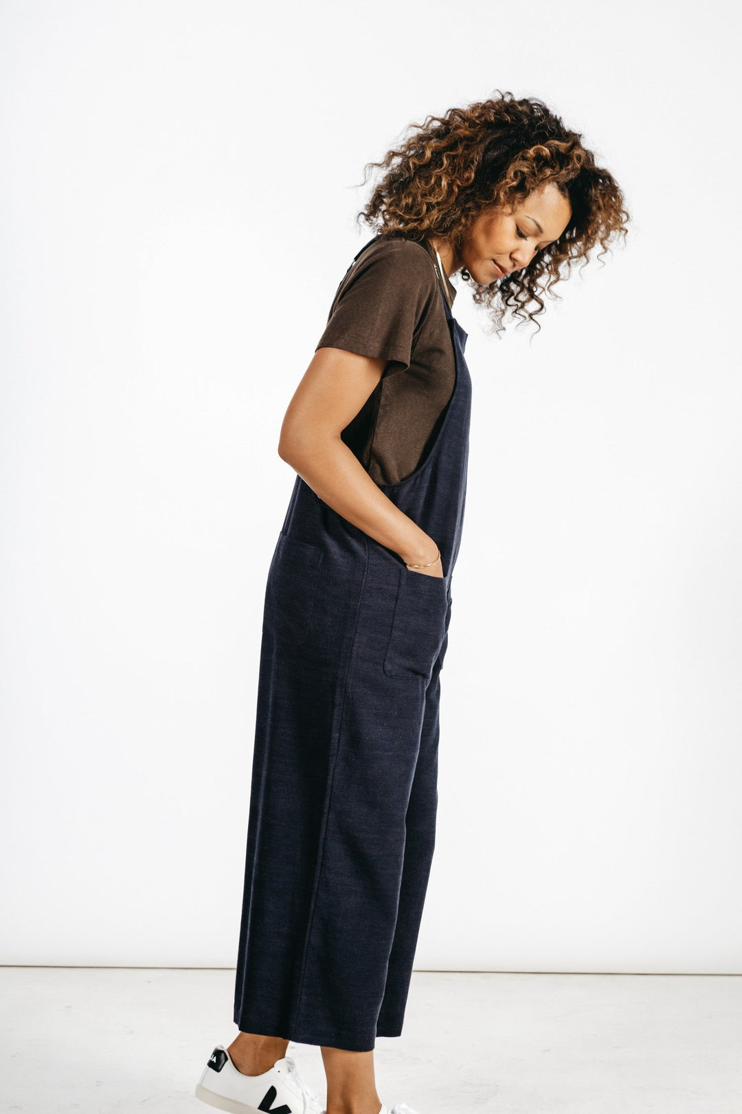 A woman with curly hair, dressed in a brown t-shirt and Bridge & Burn's Edie Overall in Navy—a comfortable linen-cotton blend style featuring adjustable straps—stands sideways with her hands in her pockets, wearing white sneakers against a plain white background.