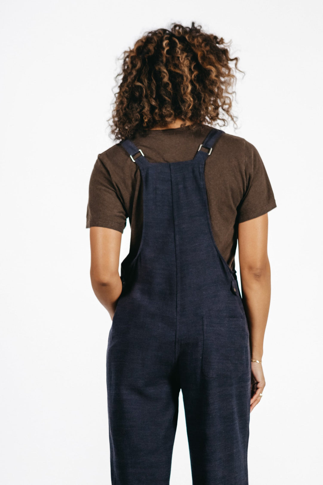 A person with curly hair, dressed in a brown t-shirt and the Bridge & Burn Edie Overall in Navy featuring adjustable straps and wide legs, stands facing away from the camera against a plain white background.