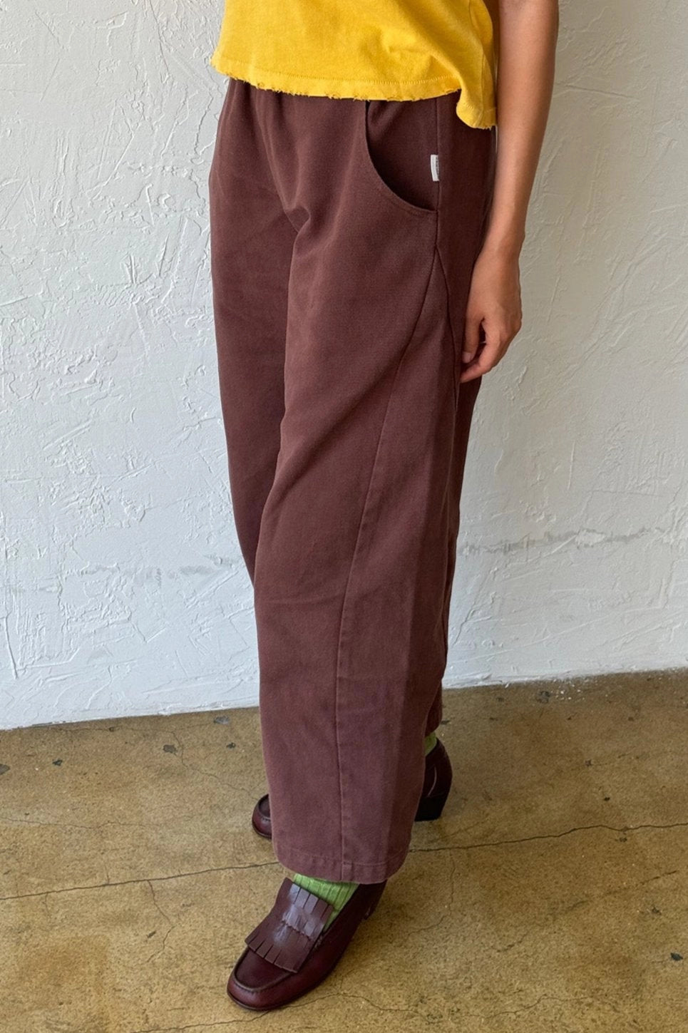 A person stands indoors against a white wall and tan floor, wearing Le Bon Shoppe - Arc Pant / Chocolate—wide-legged brown pants—paired with a yellow top, green socks, and brown loafers. Only the lower half is visible.