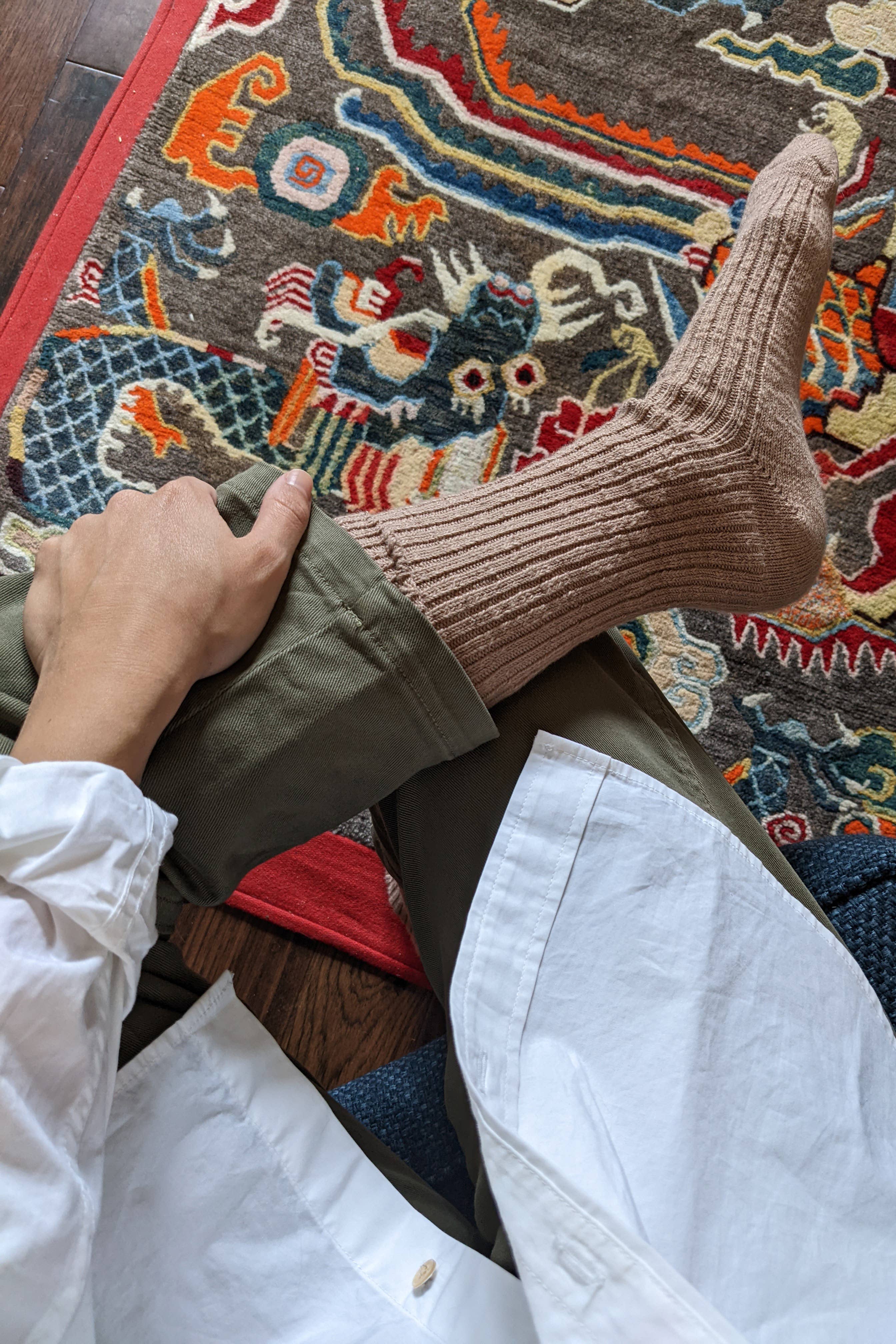A person sits cross-legged on a colorful patterned rug, wearing a white shirt, olive green pants, and Le Bon Shoppe - Cottage Socks in Toffee with their hand resting on their leg.