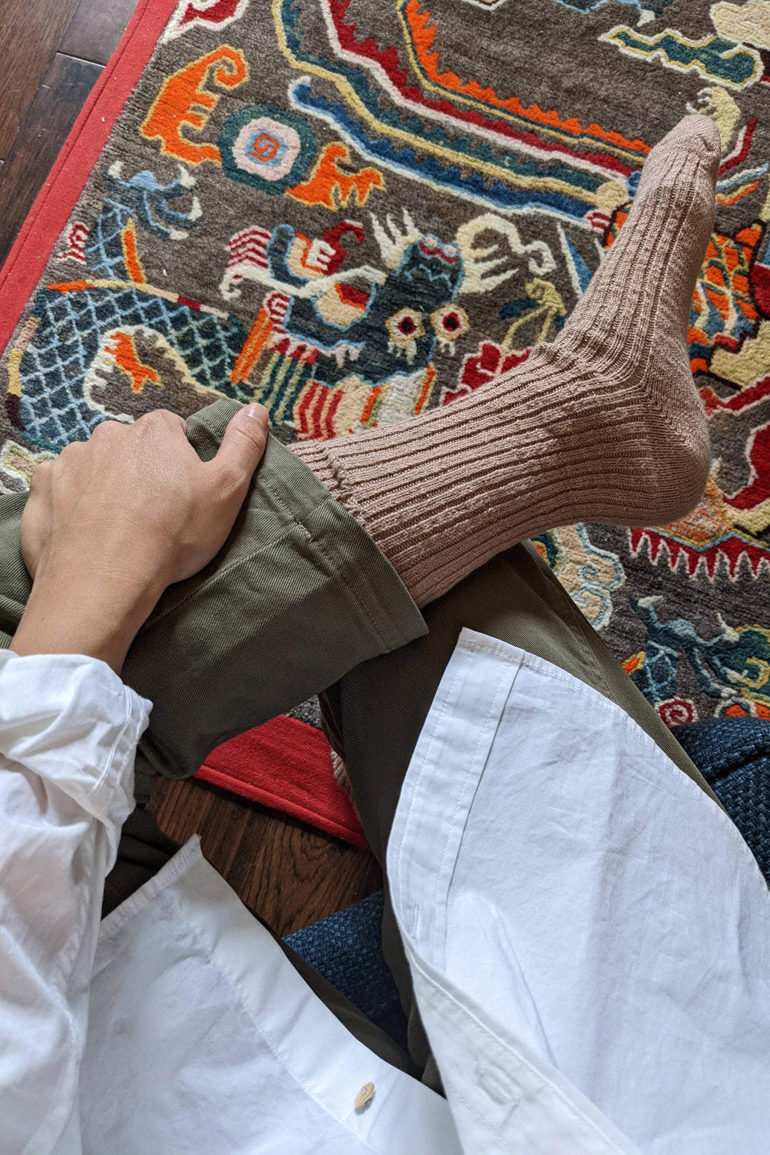 A person sits cross-legged on a colorful patterned rug, wearing a white shirt, olive green pants, and Le Bon Shoppe - Cottage Socks in Toffee with their hand resting on their leg.