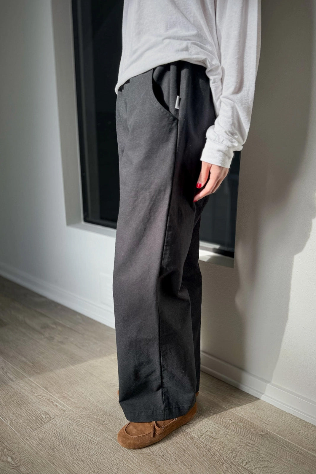 A person wearing a white long-sleeve shirt, Le Bon Shoppe - Work Pant / Black by Le Bon Shoppe, and brown slippers stands indoors on a wooden floor near a window, partially out of frame.