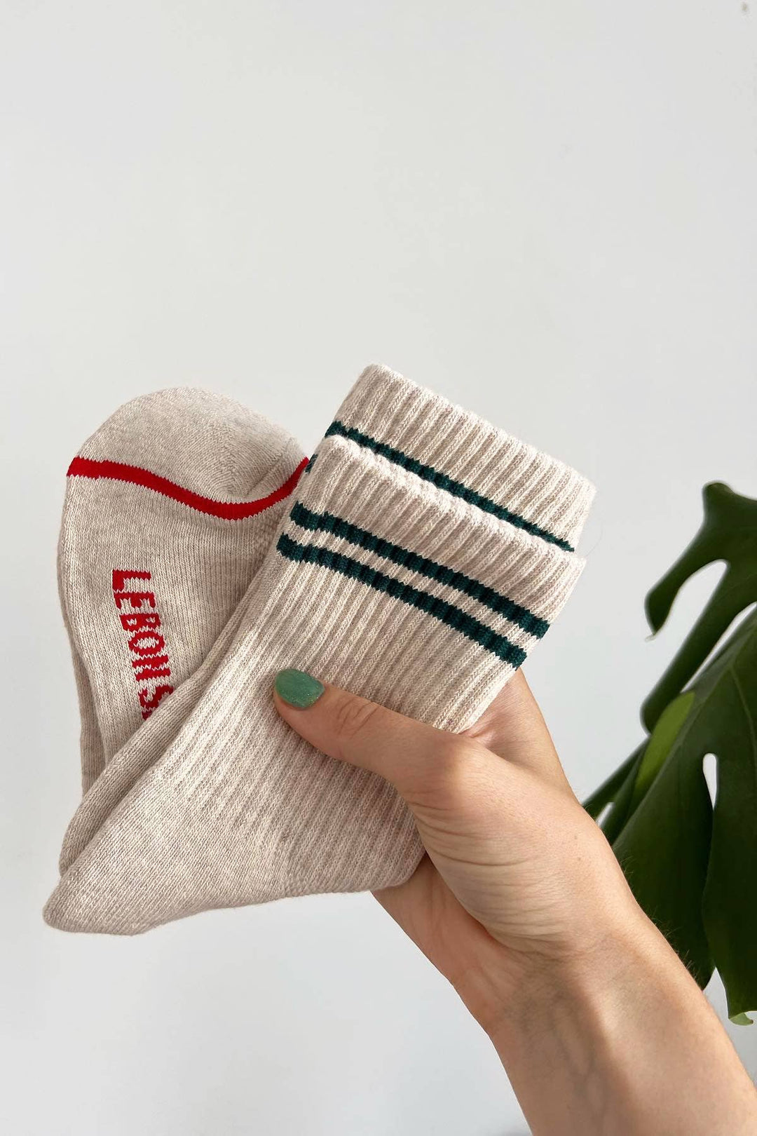 A hand with green nail polish holds Le Bon Shoppe - Boyfriend Socks in Oatmeal—one with red lettering, one with two green stripes—against a plain backdrop, accented by a green plant leaf for a cute vibe. Brand: Le Bon Shoppe.