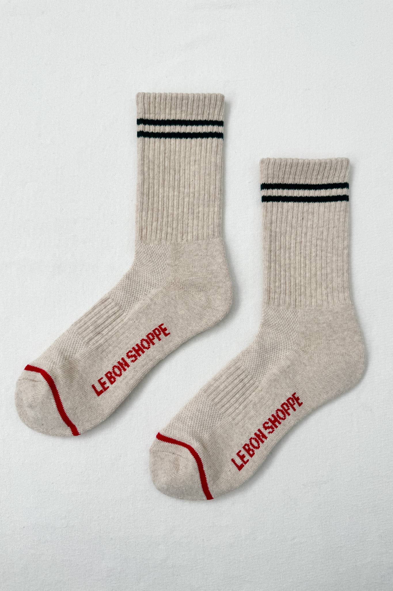 Le Bon Shoppe Boyfriend Socks in Oatmeal feature black double stripes near the top, “LE BON SHOPPE” in red on the soles, a ribbed texture, and sporty red accents at the toe and heel.