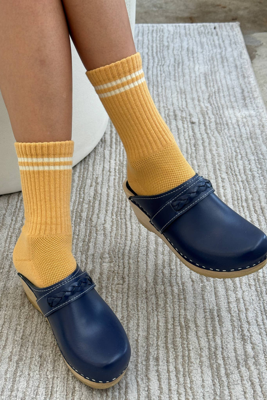A person wearing Le Bon Shoppe - Boyfriend Socks in Butter sits cross-legged on a light textured rug, pairing the yellow socks with white stripes and blue clogs.