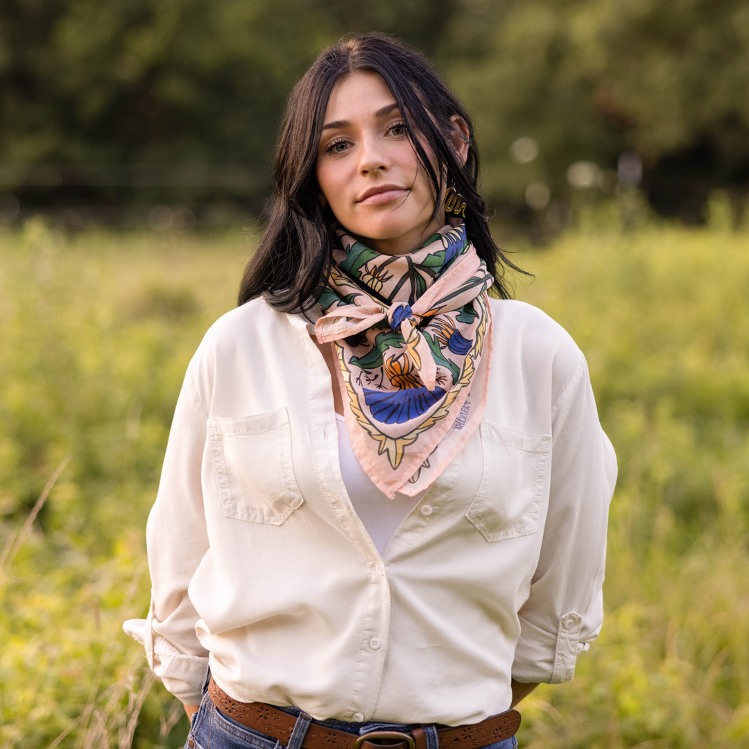 A woman with black hair stands in a grassy field, wearing a white button-up shirt and a Handker Bandanas 34" Dandy Wild Rag Bandana around her neck. She faces the camera with a neutral expression.
