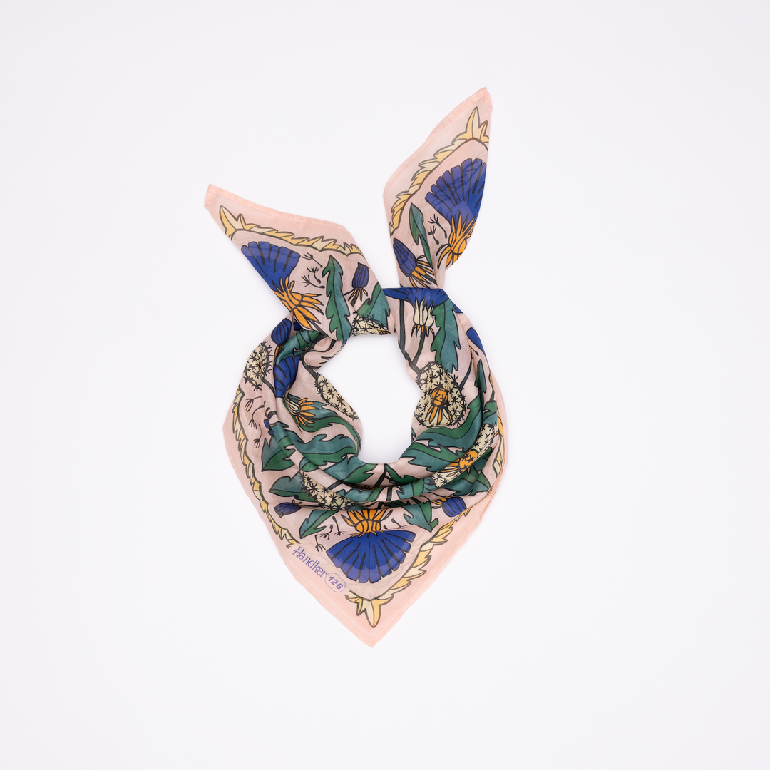 The Handker Bandanas 34" Dandy Wild Rag Bandana is a square, silk bandana with a blue, green, and yellow floral print on light pink, hand screen-printed and styled with two pointed ends facing upward.