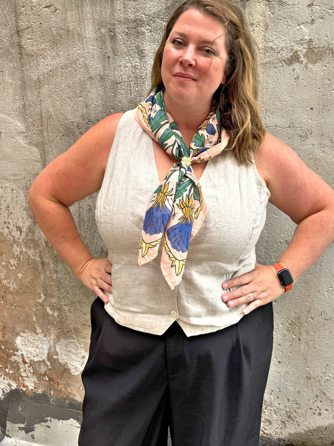 A woman poses by a weathered wall in a beige sleeveless top, black pants, and the Handker Bandanas 34" Dandy Wild Rag Bandana as a scarf. She smiles gently with her hands on her hips and wears a red smartwatch.