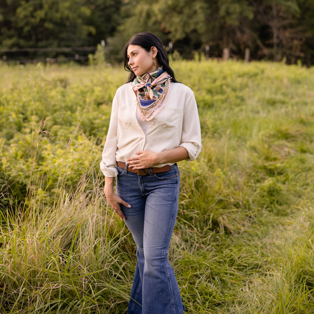 A woman stands in a grassy field, wearing a cream cotton lawn blouse, blue flared jeans, a Handker Bandanas 34" Dandy Wild Rag Bandana around her neck, and a brown belt. She gazes to the side with one hand in her pocket.