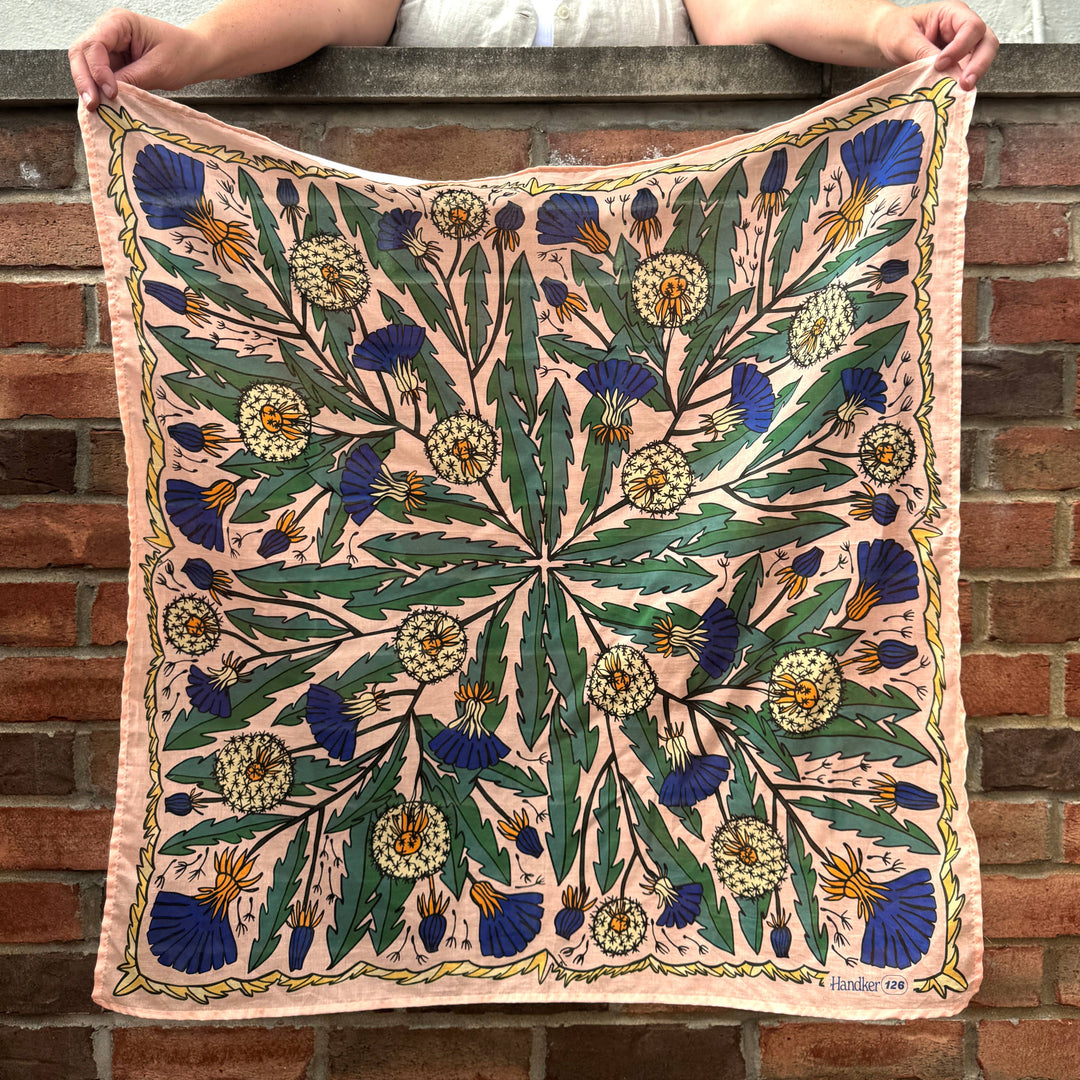 A person holds the Handker Bandanas 34" Dandy Wild Rag Bandana, made from cotton lawn with a botanical print of green leaves, blue and yellow flowers, and beige dandelions on peach, standing in front of a brick wall.