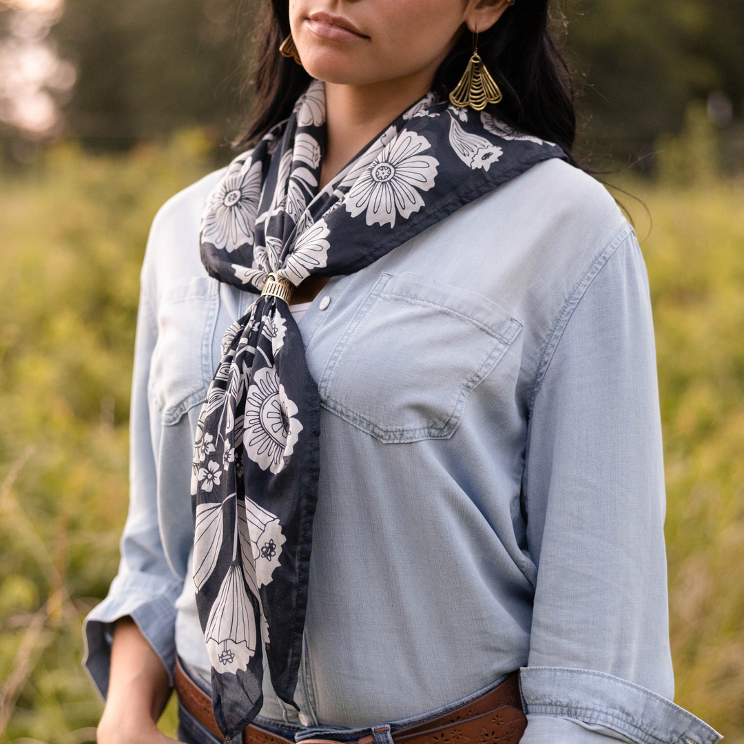 A woman with long dark hair and gold fan earrings stands outdoors in a light denim shirt, brown belt, and a Handker Bandanas 34" Wild Rag Fiona Bandana worn as a dark floral scarf. The green background is softly blurred.