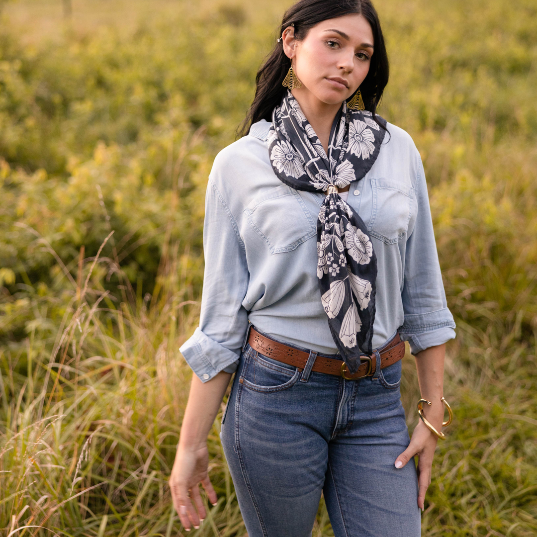 A woman stands in a grassy field wearing a Handker Bandanas 34" Wild Rag Fiona Bandana with a white floral pattern, paired with a light denim shirt and blue jeans. She has long dark hair, gold earrings, and looks calmly at the camera.