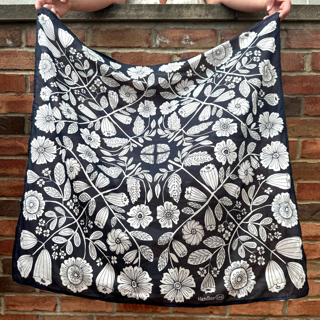 A person holds up the Handker Bandanas 34" Wild Rag Fiona Bandana in black cotton lawn, featuring a white, hand screen-printed floral design in an intricate, symmetrical pattern against a brick wall backdrop.