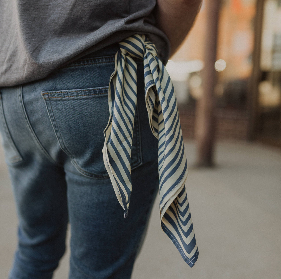 A person in blue jeans and a gray shirt wears the Handker Bandanas - No. 015 Sammie Bandana by Handker Bandanas, tied at their waist with ends hanging down, set against an outdoor urban backdrop.