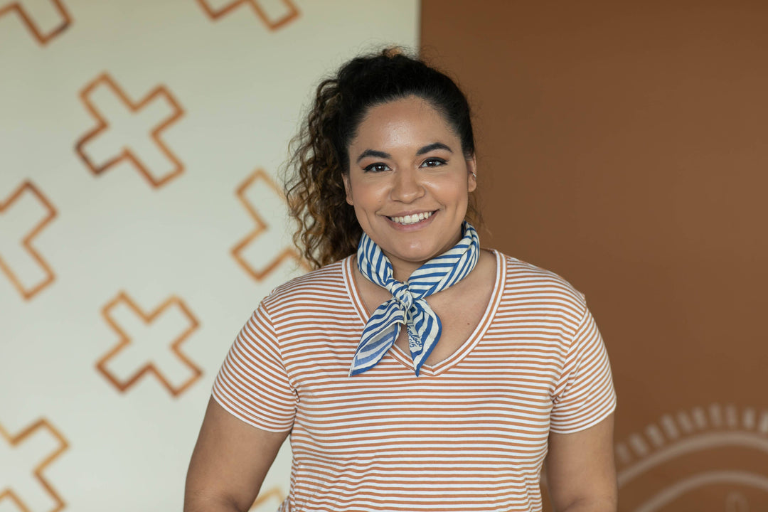 A smiling woman with curly hair in a ponytail wears a striped shirt and the Handker Bandanas - No. 015 Sammie Bandana, a blue-and-white, hand screen-printed cotton accessory (22x22 in), standing before geometric white and brown patterns.