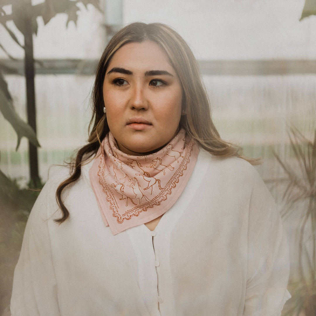 A woman with long, light brown hair wears the Handker Bandanas - No. 062 Horses Bandana by Handker Bandanas—a premium cotton bandana with embroidered details. She stands indoors near green plants, gazing to the side with a neutral expression.