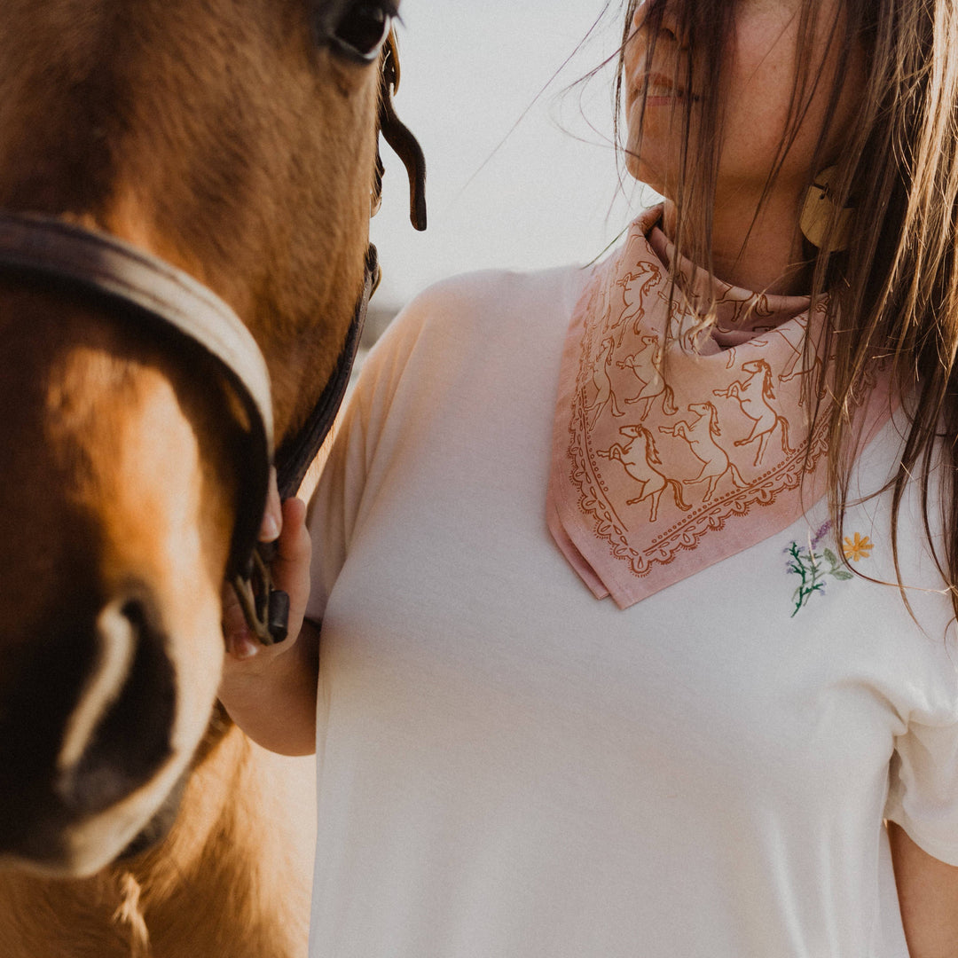 A woman in a white shirt with floral embroidery wears the Handker Bandanas - No. 062 Horses Bandana by Handker Bandanas as she stands beside a brown horse, gently holding its bridle. Her face is partially out of frame.