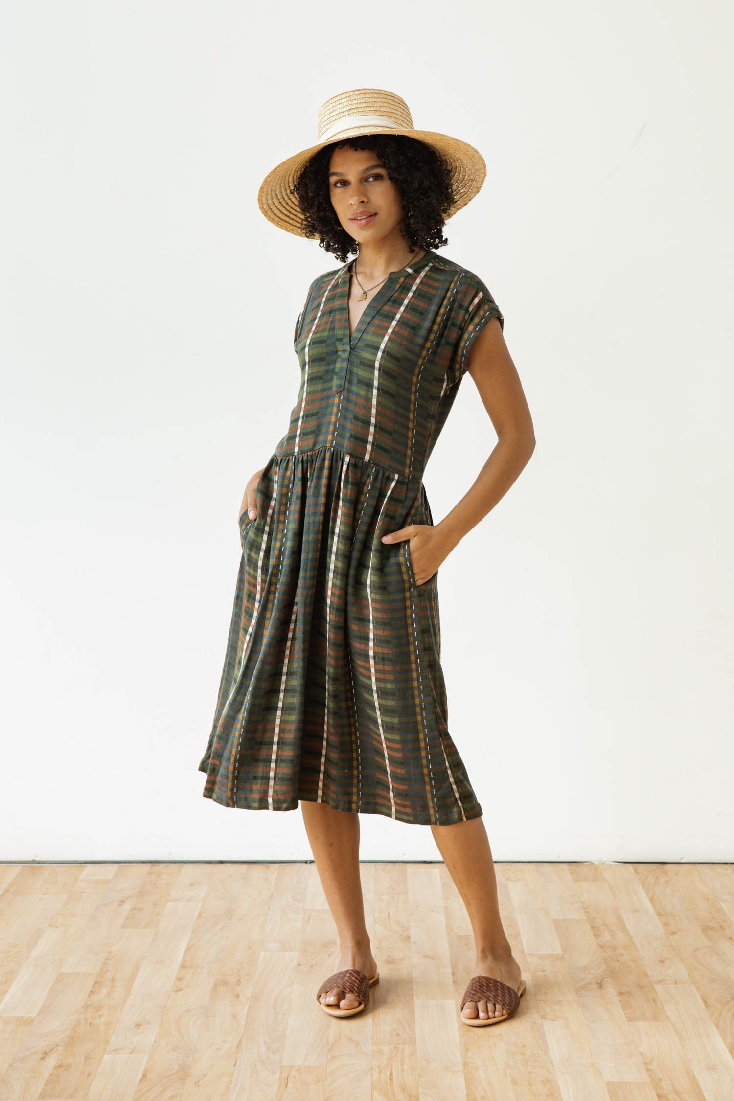 The Bridge & Burn Ira Dress in Olive Stripe is a short-sleeve, knee-length pullover dress with a dark multicolored striped pattern, featuring a gathered waist and split neckline. Crafted from a soft modal-linen blend, it is shown laid flat on a white background.