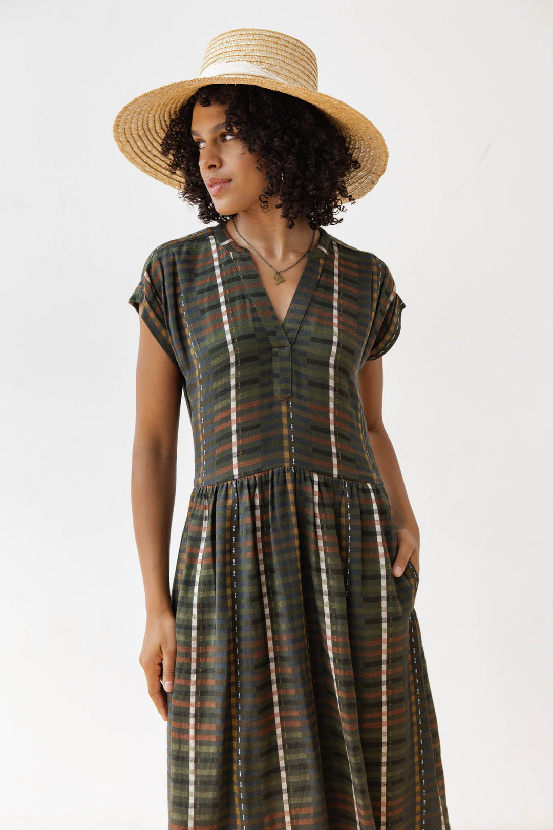 A woman in the Bridge & Burn Ira Dress in Olive Stripe, a wrinkle-resistant pullover with short sleeves, stands against a white background wearing a wide-brimmed straw hat, looking to the side with one hand in her pocket.