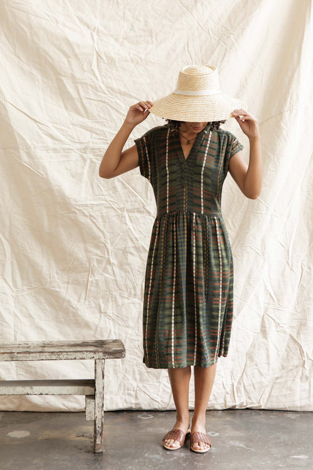 Wearing the Bridge & Burn Ira Dress in Olive Stripe, a person stands indoors on a gray floor, adjusting a wide-brimmed straw hat next to a rustic wooden bench and a light, wrinkled fabric backdrop.