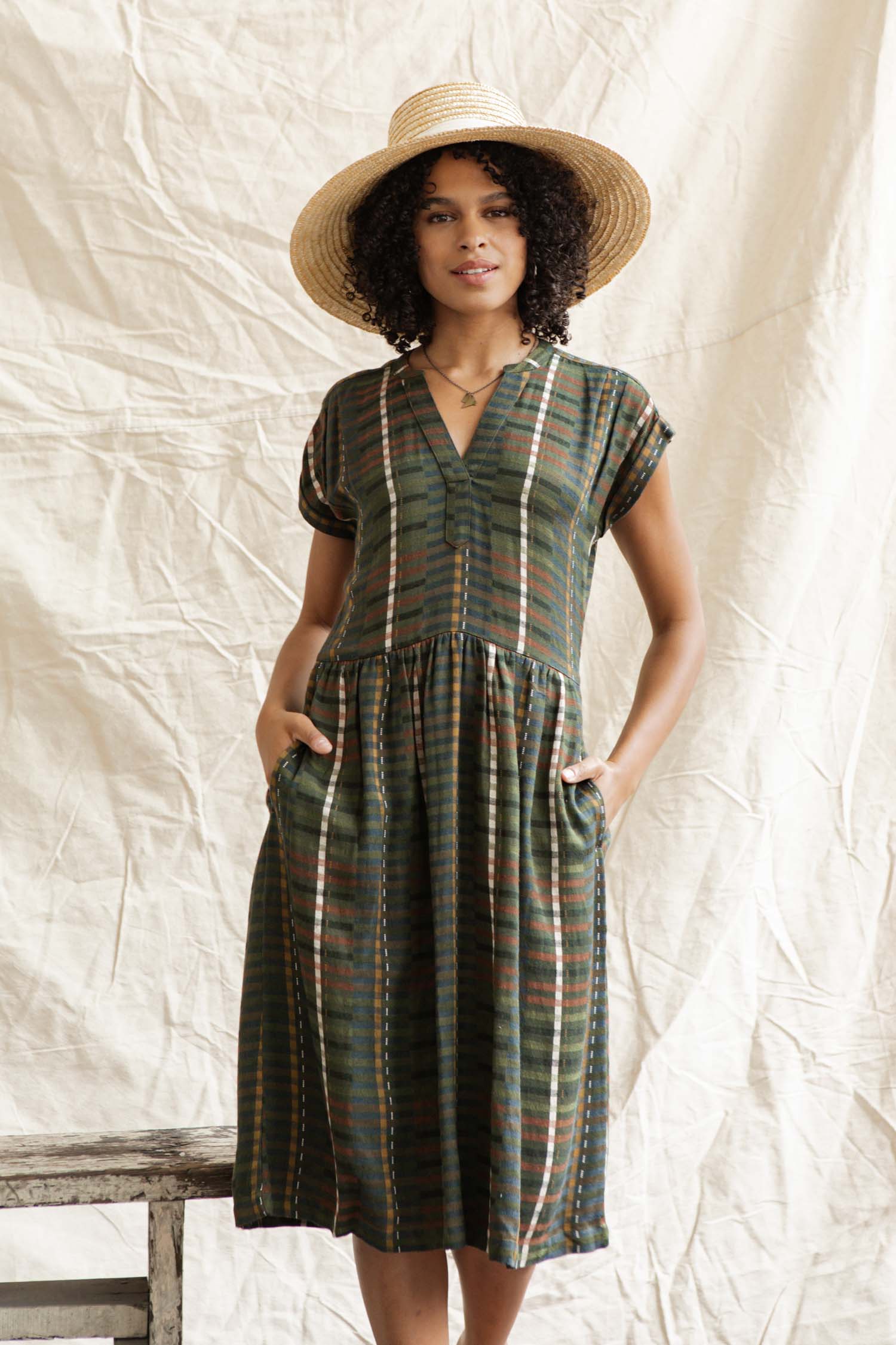 A woman with curly hair and a wide-brimmed straw hat stands before a cream backdrop, wearing the Bridge & Burn Ira Dress in Olive Stripe—a short-sleeve pullover dress with pockets.