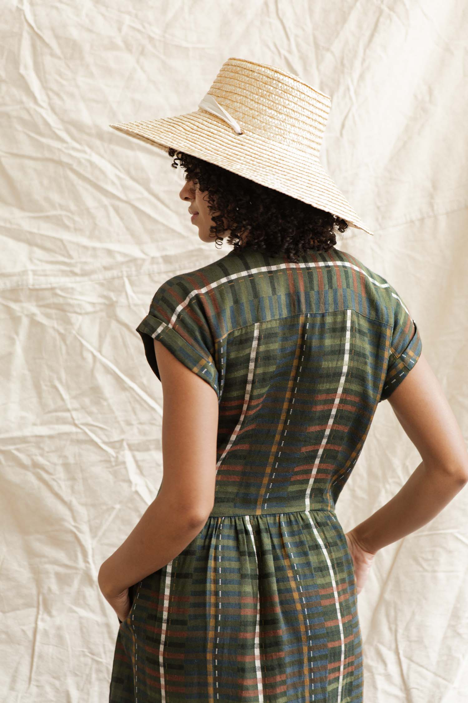 A person with curly hair wears the Bridge & Burn Ira Dress in Olive Stripe and a wide-brimmed straw hat, standing with their back turned against a light, textured fabric backdrop.