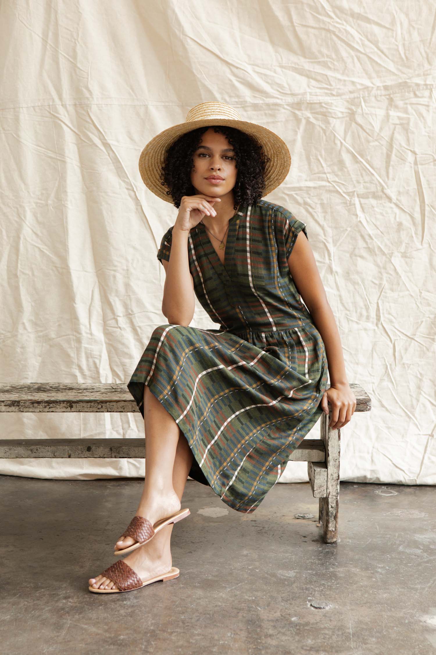 A woman in the Bridge & Burn Ira Dress in Olive Stripe and brown sandals sits on a rustic bench against a beige fabric backdrop, resting her chin on her hand beneath a wide-brimmed straw hat, gazing at the camera.