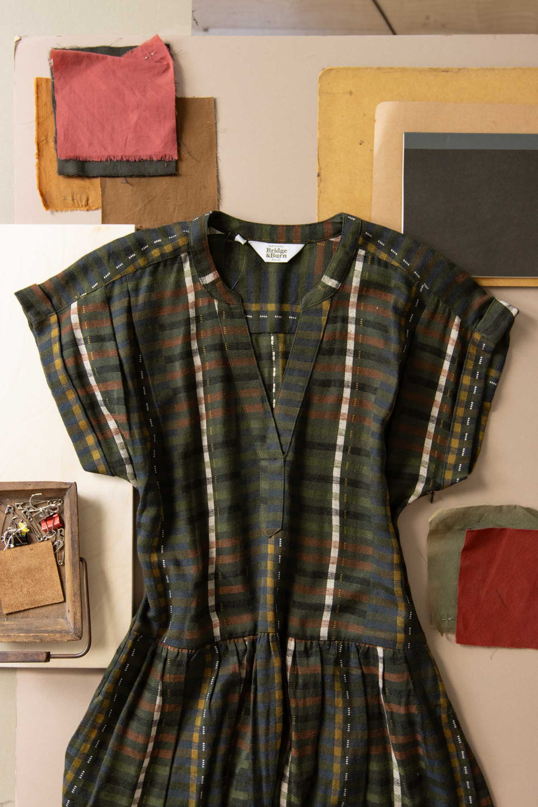 The Ira Dress in Olive Stripe by Bridge & Burn, a short-sleeved modal linen piece, lies flat among fabric swatches, papers, and a small tray of sewing supplies.
