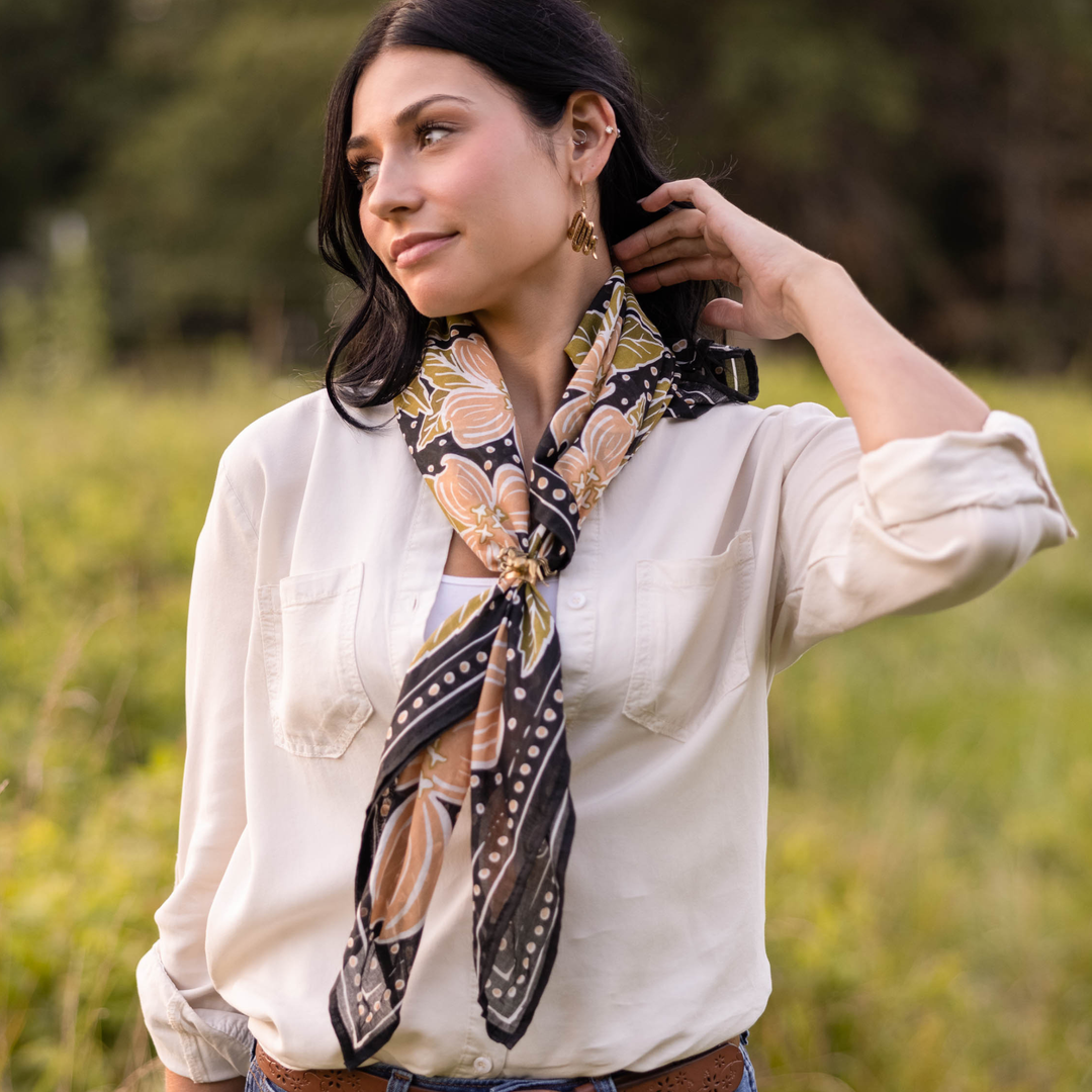 A woman with long dark hair stands in a grassy field, wearing a white button-up shirt and the Handker Bandanas 34" Wild Rag Amy Bandana around her neck. She looks to the side, one hand touching her neck.