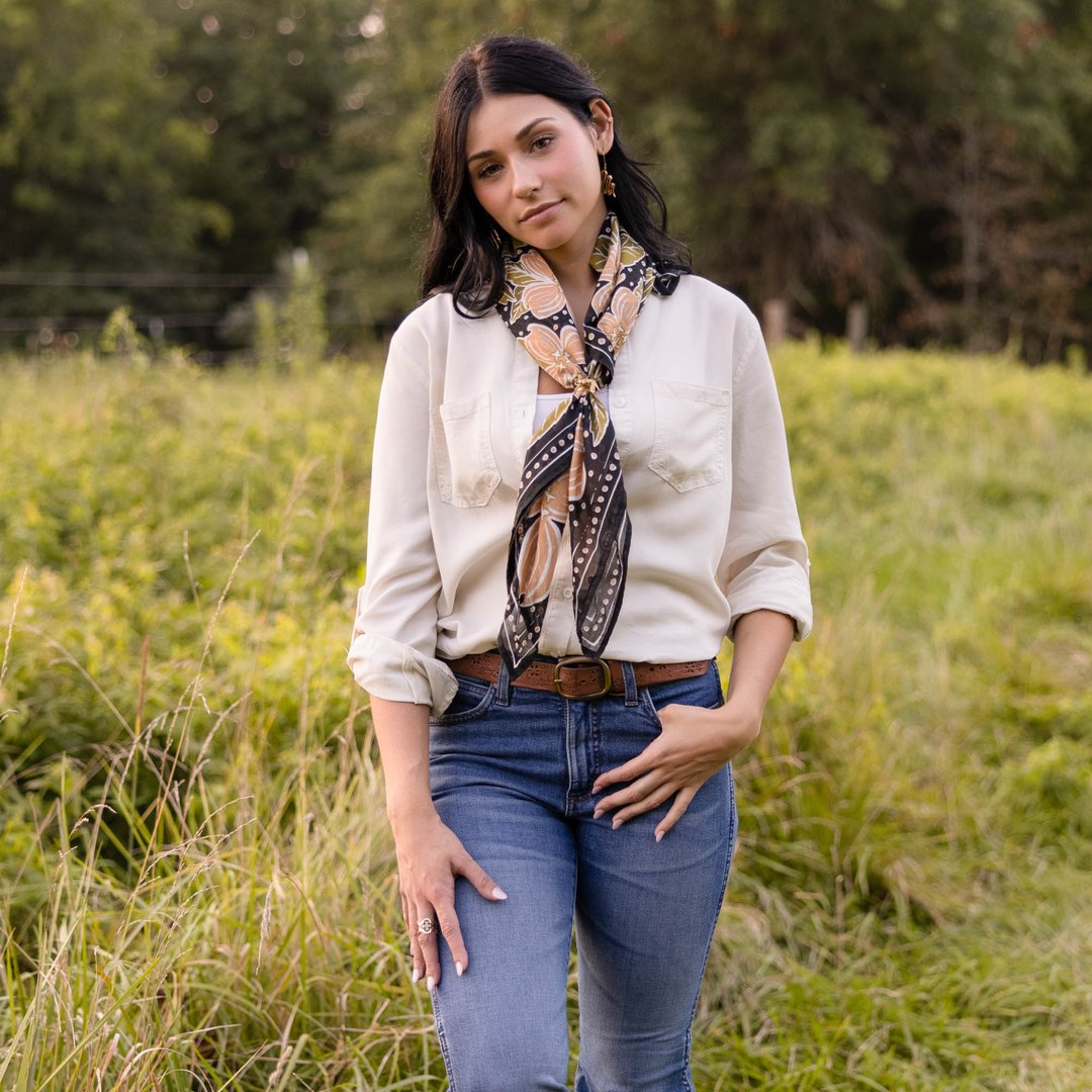 A woman with dark hair stands in a grassy field, wearing blue jeans, a cream blouse, and the Handker Bandanas 34" Wild Rag Amy Bandana around her neck. She looks thoughtful with one hand in her pocket; trees fill the background.