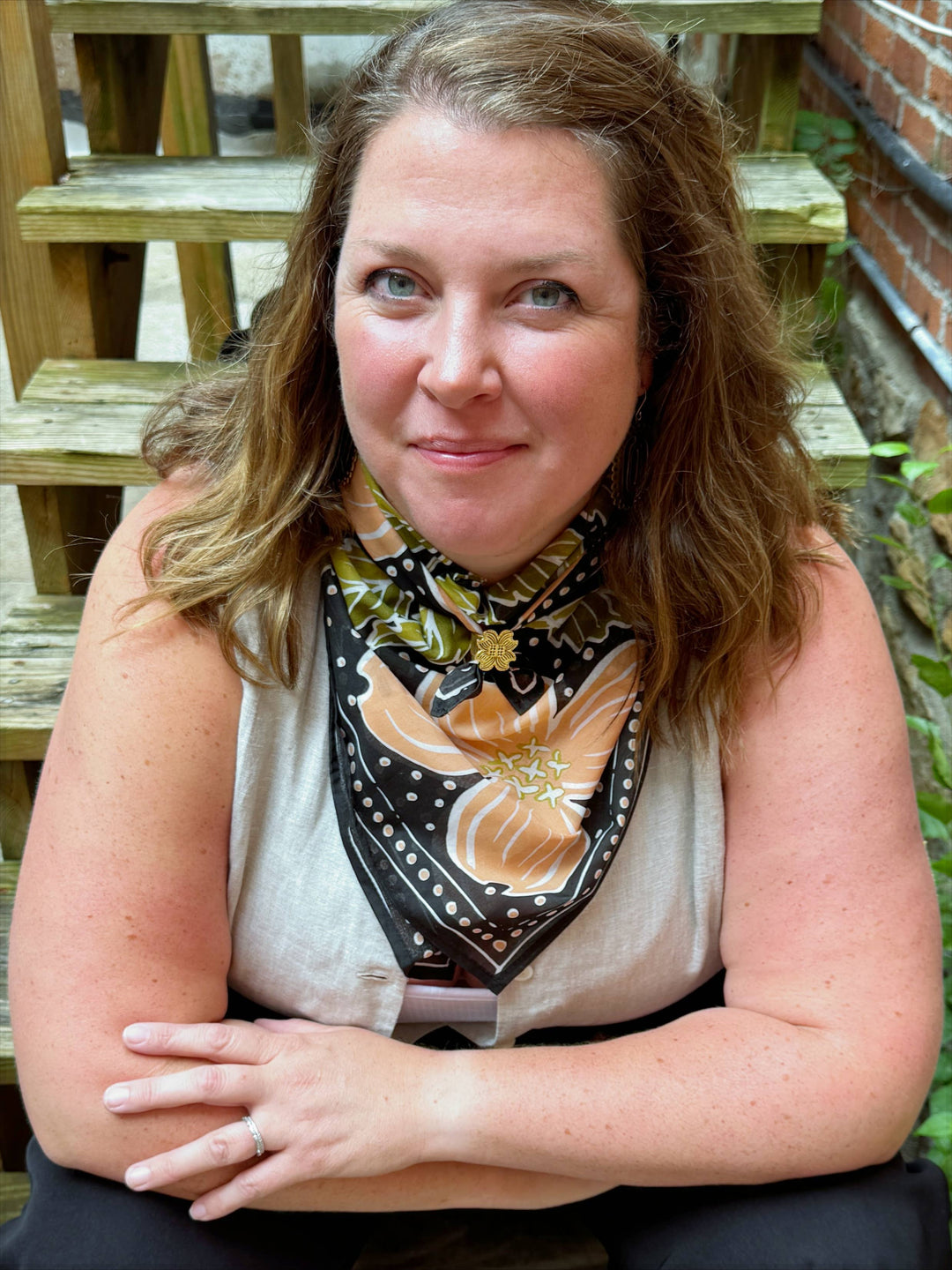 A woman with wavy brown hair sits on wooden steps, smiling slightly at the camera while wearing the Handker Bandanas 34" Wild Rag Amy Bandana and a light sleeveless top. Brick and greenery appear in the background.