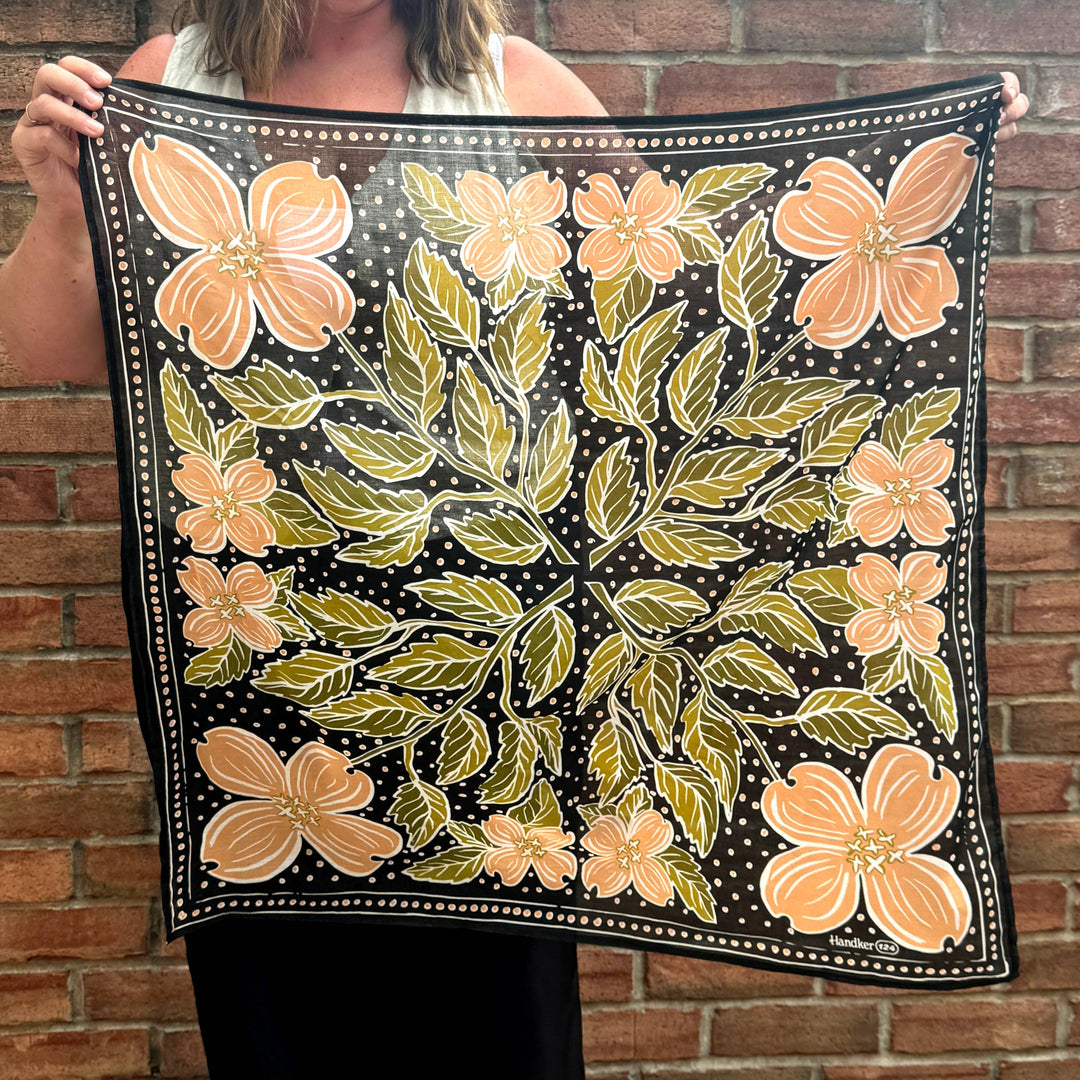 A person holds the Handker Bandanas 34" Wild Rag Amy Bandana, featuring a black background with orange flowers, green leaves, and small white dots, while standing in front of a brick wall.