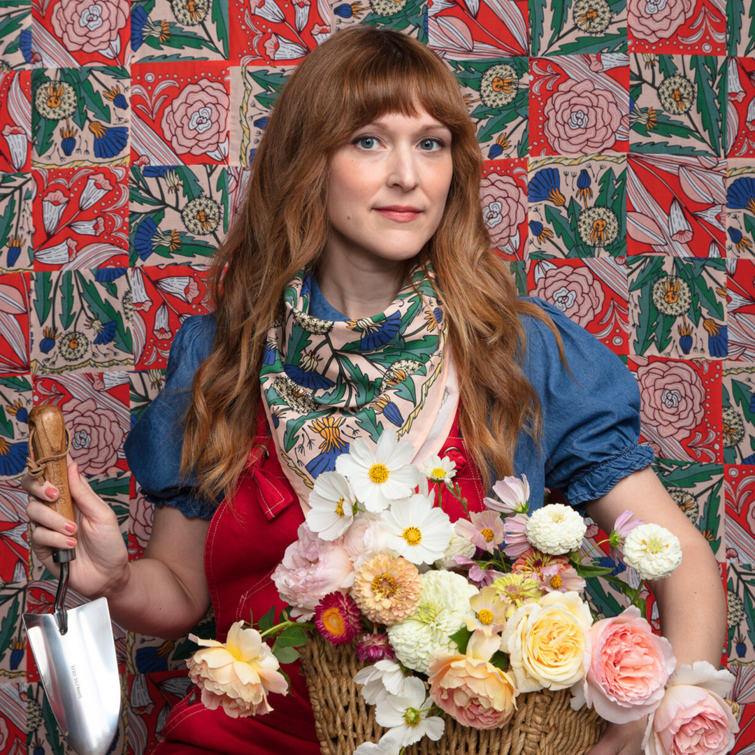 A woman with long auburn hair wears a blue shirt, red apron, and the Handker Bandanas No. 103 Dandy Bandana while holding a garden trowel and basket of flowers against a vibrant floral-patterned background.