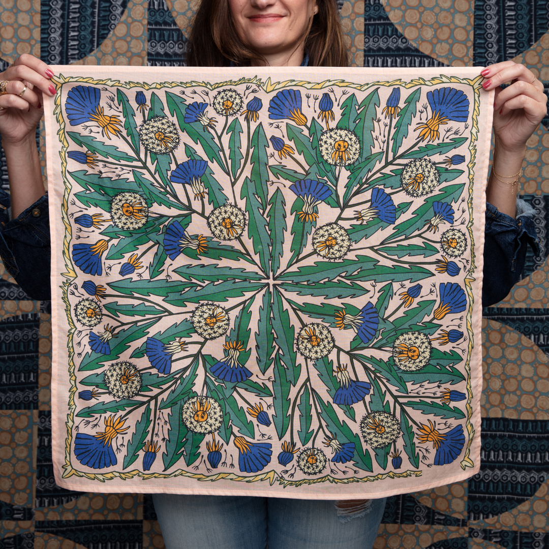 A woman holds up the Handker Bandanas - No. 103 Dandy Bandana by Handker Bandanas, a premium cotton bandana with a symmetrical botanical design of green leaves and blue and yellow flowers, against a patterned background.