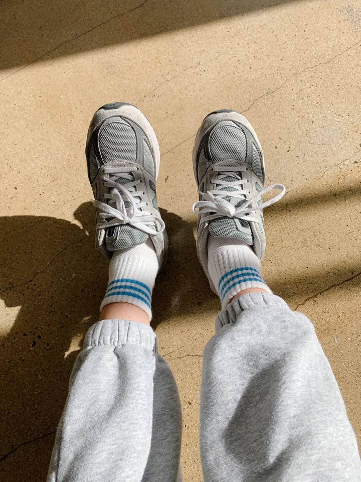 A person wearing grey sneakers, Le Bon Shoppe - Girlfriend Socks in Ivory, and light grey sweatpants stands on a sunlit beige floor.