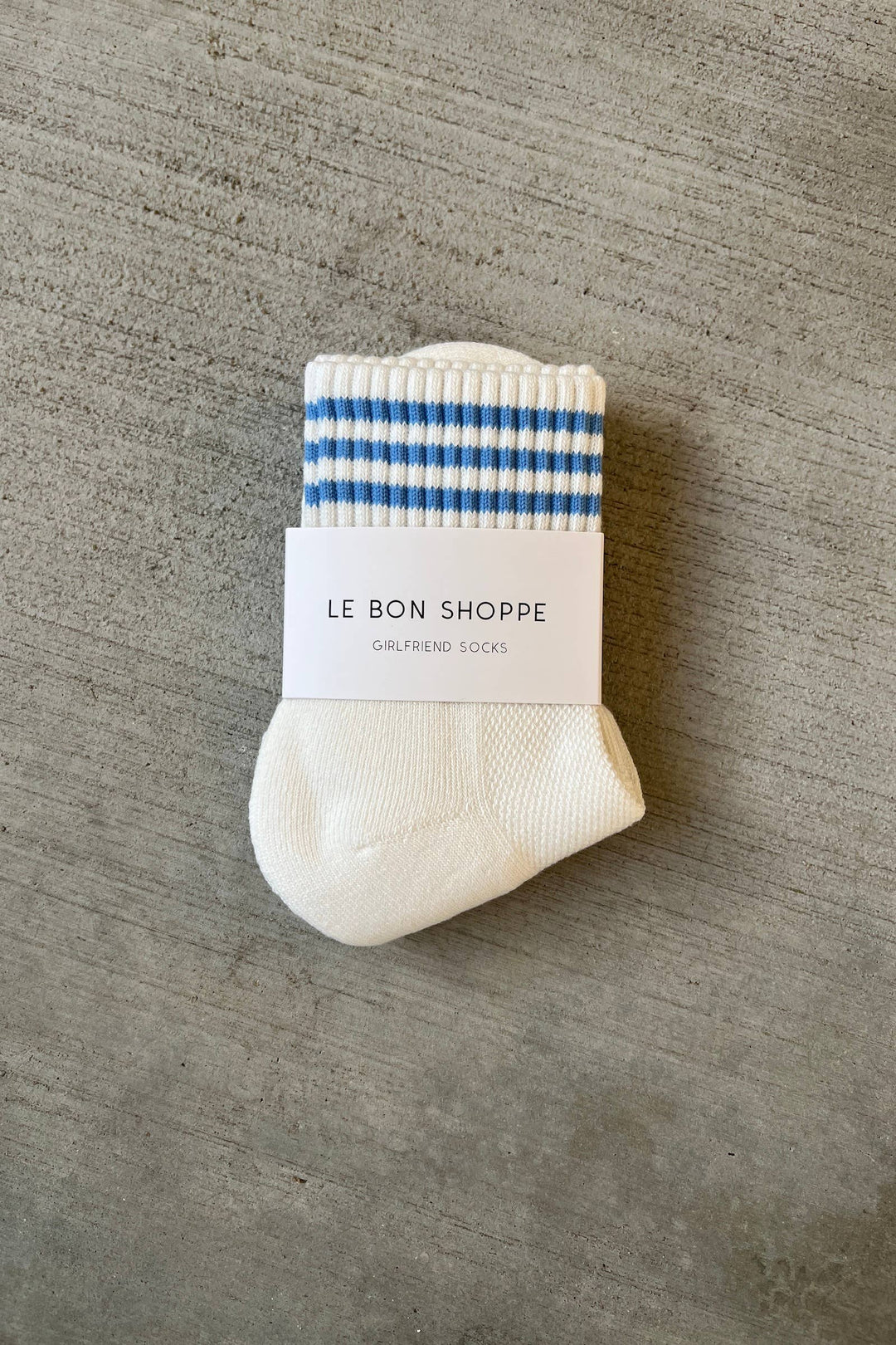 A pair of Le Bon Shoppe - Girlfriend Socks in Ivory, featuring blue and white stripes at the top, is folded with its branded label and displayed on a textured gray surface—ideal for everyday comfort.