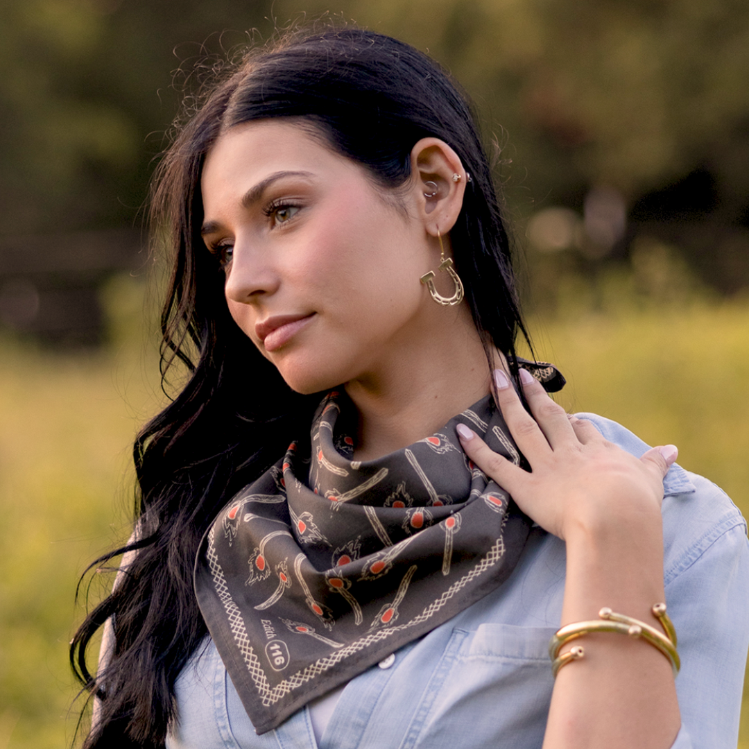 A woman with long dark hair wears the Handker Bandanas - No. 116 Edith Bandana in charcoal gray by Handker Bandanas, paired with gold jewelry and a light blue shirt, standing outdoors and gazing serenely to the side.